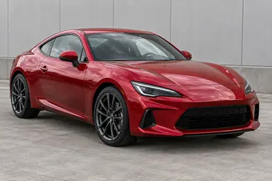 Deep-red sports coupe beside an industrial wall with soft reflections across its hood.