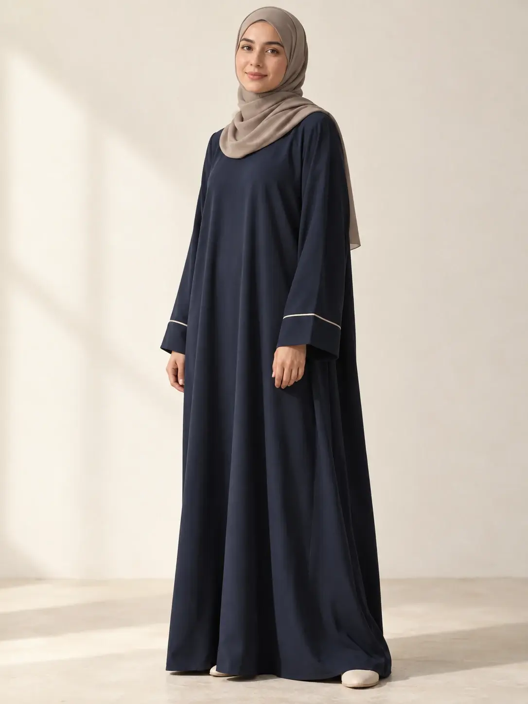 Woman wearing a navy abaya with cream cuff piping and a taupe headscarf in an off-white studio.