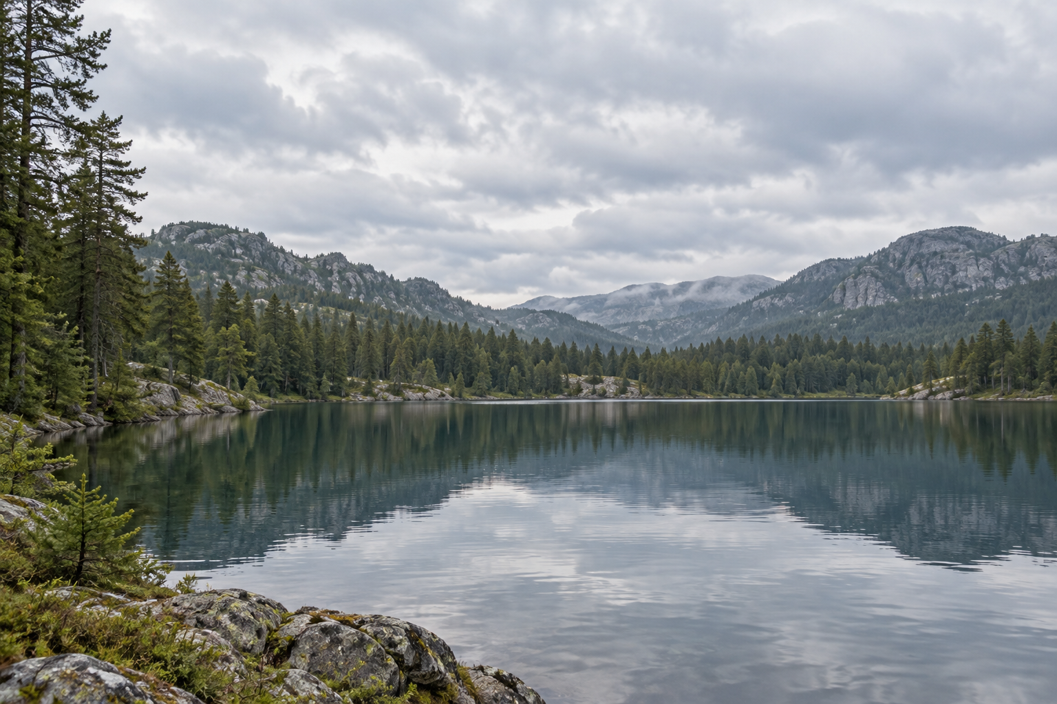 Mountain lake and pine trees beneath layered overcast clouds reflected in calm water.